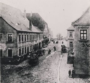 Historische Schwarz-Weiß-Aufnahme der gepflasterten Brückenstraße in Haßfurt um 1900 mit Passanten und Fuhrwerken; links angeschnitten das Gasthaus „Zum Goldenen Löwen“, rechts Wohn- und Geschäftshäuser.