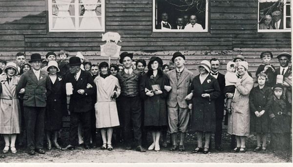 Gruppenfoto von der Einweihungsfeier des FC-Sportheims 1931: zahlreiche festlich gekleidete Männer, Frauen und Kinder mit Hüten und Mänteln stehen vor der Holzwand des neuen Sportheims, in der Mitte wird ein Schild hochgehalten, aus den Fenstern schauen weitere Gäste.