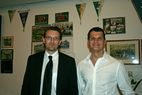 Zwei Männer in Anzug und weißem Hemd vor einer Wand mit Fotos und Wimpeln