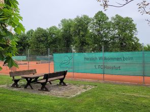 Sitzgruppe an der Tennisanlage, am Zaun ein Banner: Herzlich Willkommen beim 1. FC Haßfurt