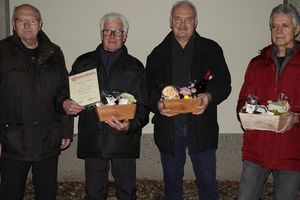 Vier Männer in Winterjacken vor einer hellen Wand; der zweite von links hält eine Ehrenurkunde und einen Geschenkkorb, die beiden rechts halten ebenfalls Geschenkkörbe