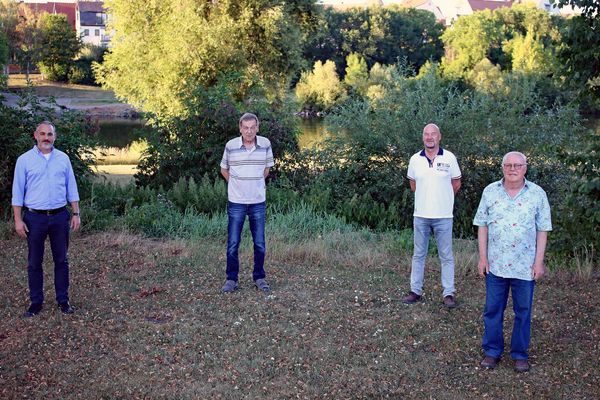 Vier Männer stehen mit großem Abstand nebeneinander auf einer Wiese, im Hintergrund Büsche, Bäume, ein Flusslauf und Häuser
