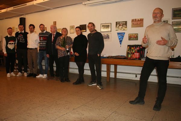 Gruppe im Vereinsheim vor einer Wand mit gerahmten Fotos, ein Mann spricht zu den Anwesenden