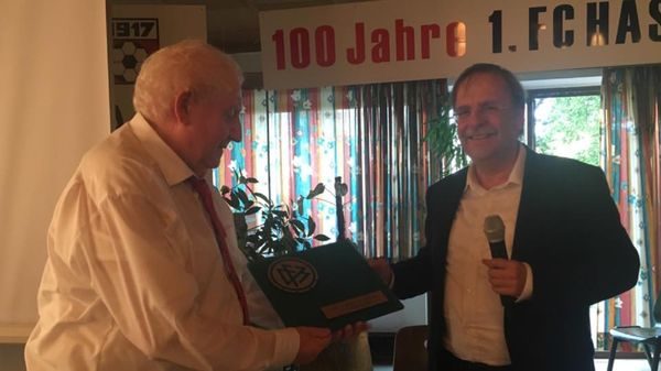 Redner mit Mikrofon übergibt einem älteren Herrn eine Mappe mit DFB-Logo vor dem Banner „100 Jahre 1. FC Haßfurt“