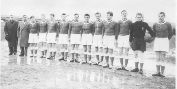 Schwarz-Weiß-Foto der Meistermannschaft vom 24.02.1957: elf Spieler in dunklen Trikots mit hellem Kragen und weißen Hosen stehen in einer Reihe auf einem nassen, schlammigen Platz, der Torwart im dunklen Pullover hält einen Ball; links zwei Männer in Mänteln, im Hintergrund eine Brücke.