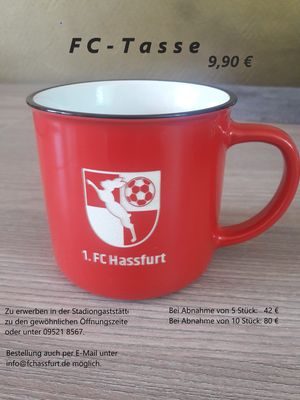 Rote Kaffeetasse mit weißem Vereinswappen und Schriftzug „1. FC Hassfurt“ auf einem Holztisch, Angebotsplakat „FC-Tasse 9,90 €“ mit Mengenpreisen
