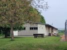Tennisheim von außen: heller Flachbau mit Fensterreihe, davor Rasen, links ein Baum und eine Sitzbank
