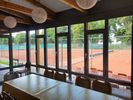 Blick aus dem Gastraum des Tennisheims durch die bodentiefe Fensterfront auf die Sandplätze